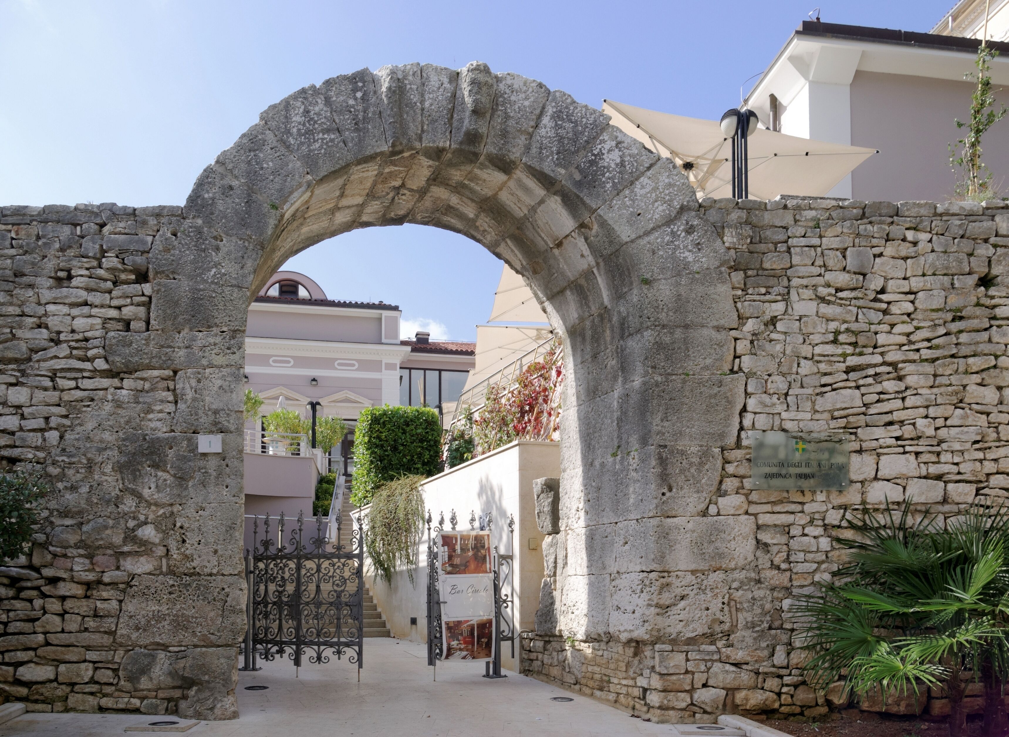 An ancient stone archway leading to a courtyard with a gate, surrounded by stone walls and vegetation.