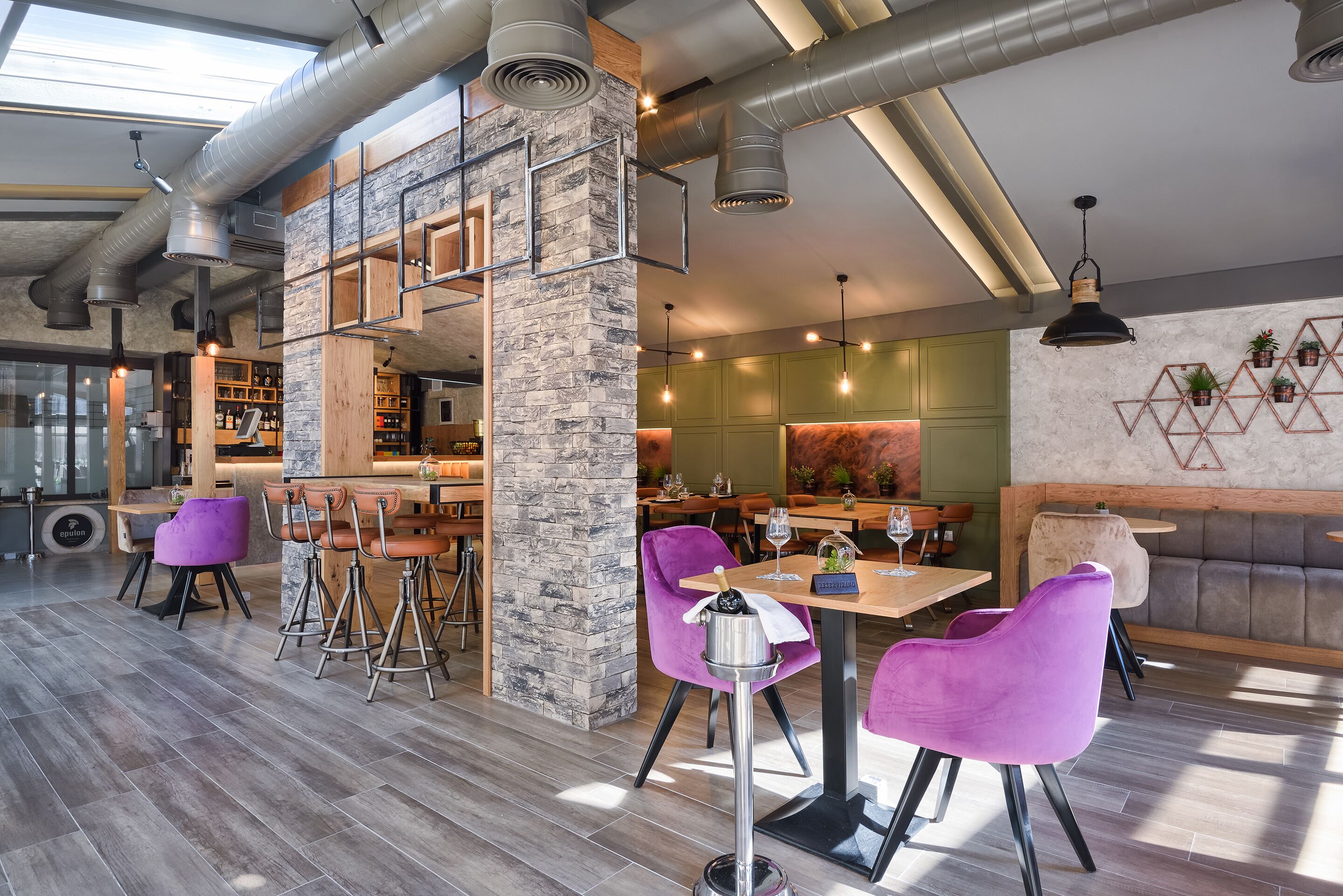 Un restaurant moderne avec un plafond haut, des conduits apparents et un mélange d’éléments en bois et en pierre. On y trouve des chaises violettes et un coin bar avec des tabourets.