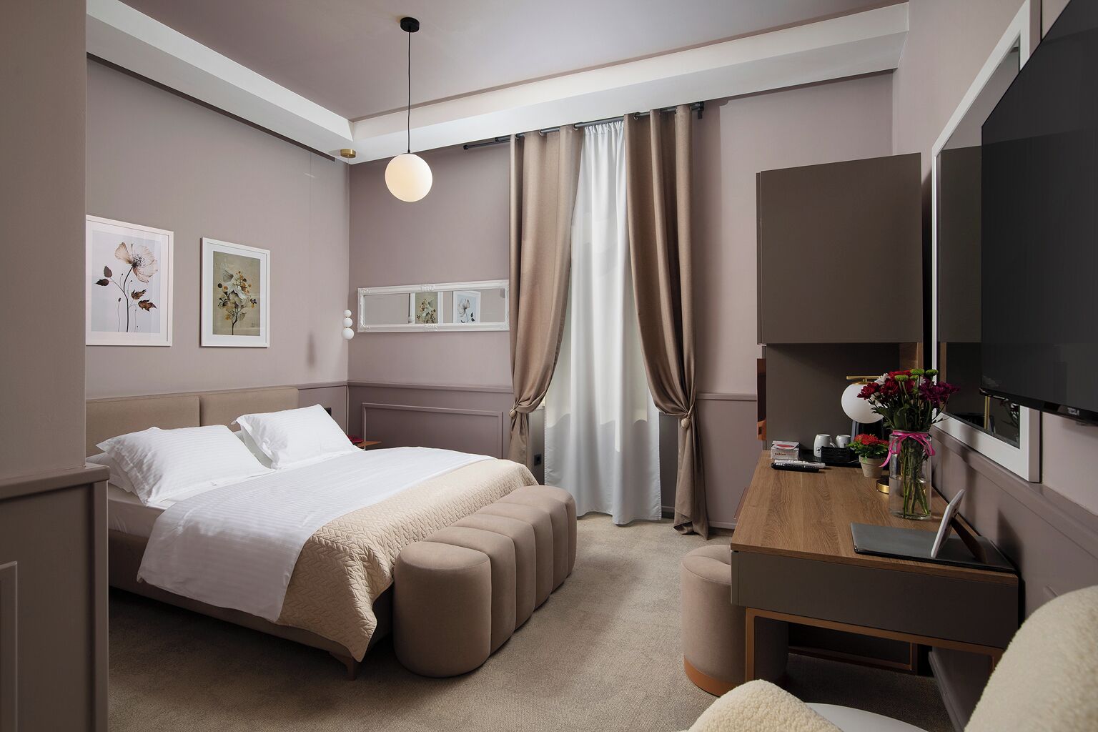 A hotel room with a bed, a desk with a TV, a chair, a bedside table, and a window with curtains.