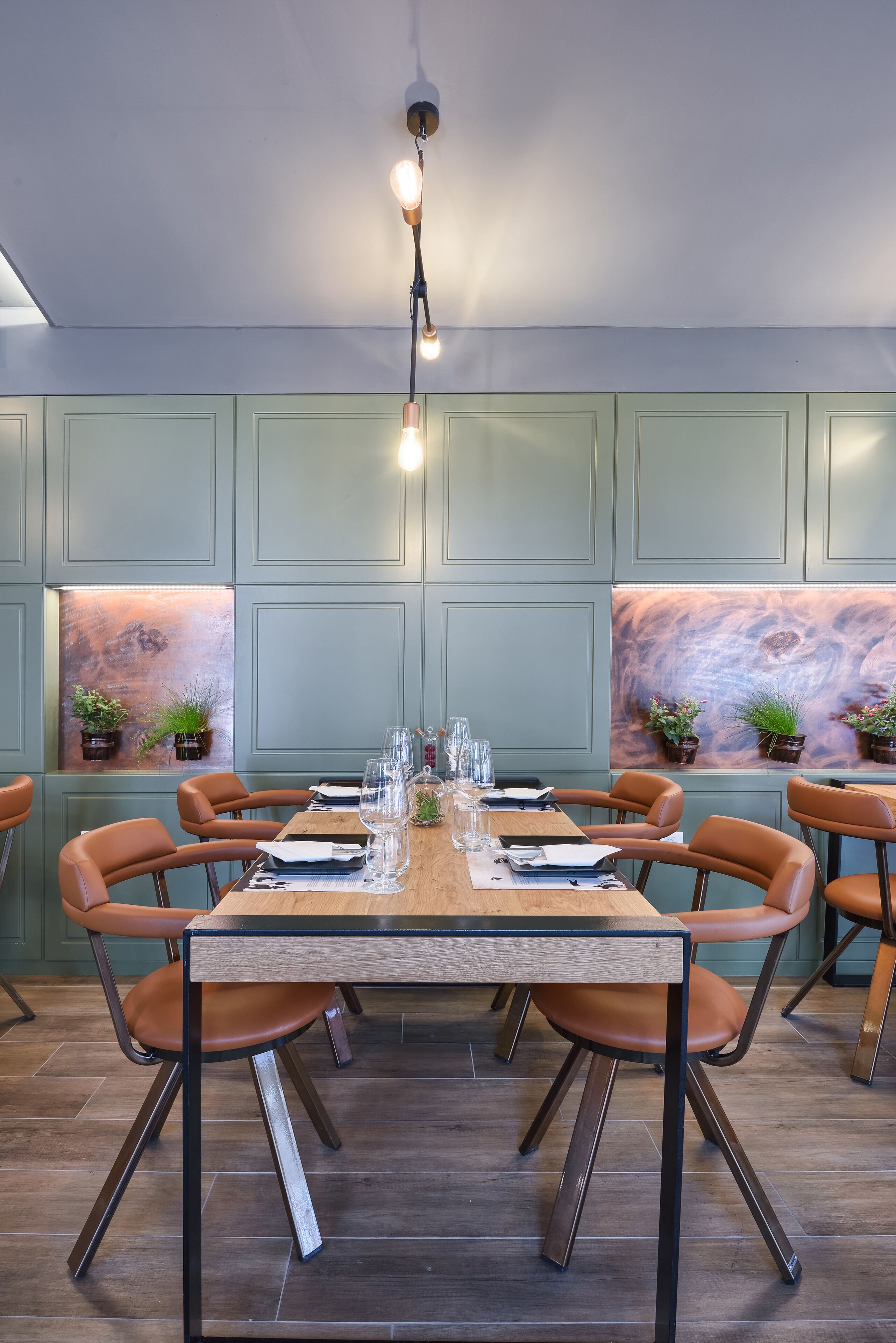 A dining area with a long wooden table set for six, surrounded by six brown leather chairs. The walls are painted in a light teal color, and there are two windows with potted plants on either side.
