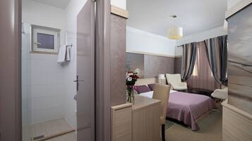 Ein Hotelzimmer mit Dusche, einem Bett mit violettem Überwurf, einem Schreibtisch mit Blumen in einer Vase und Blick ins Freie durch das Fenster.