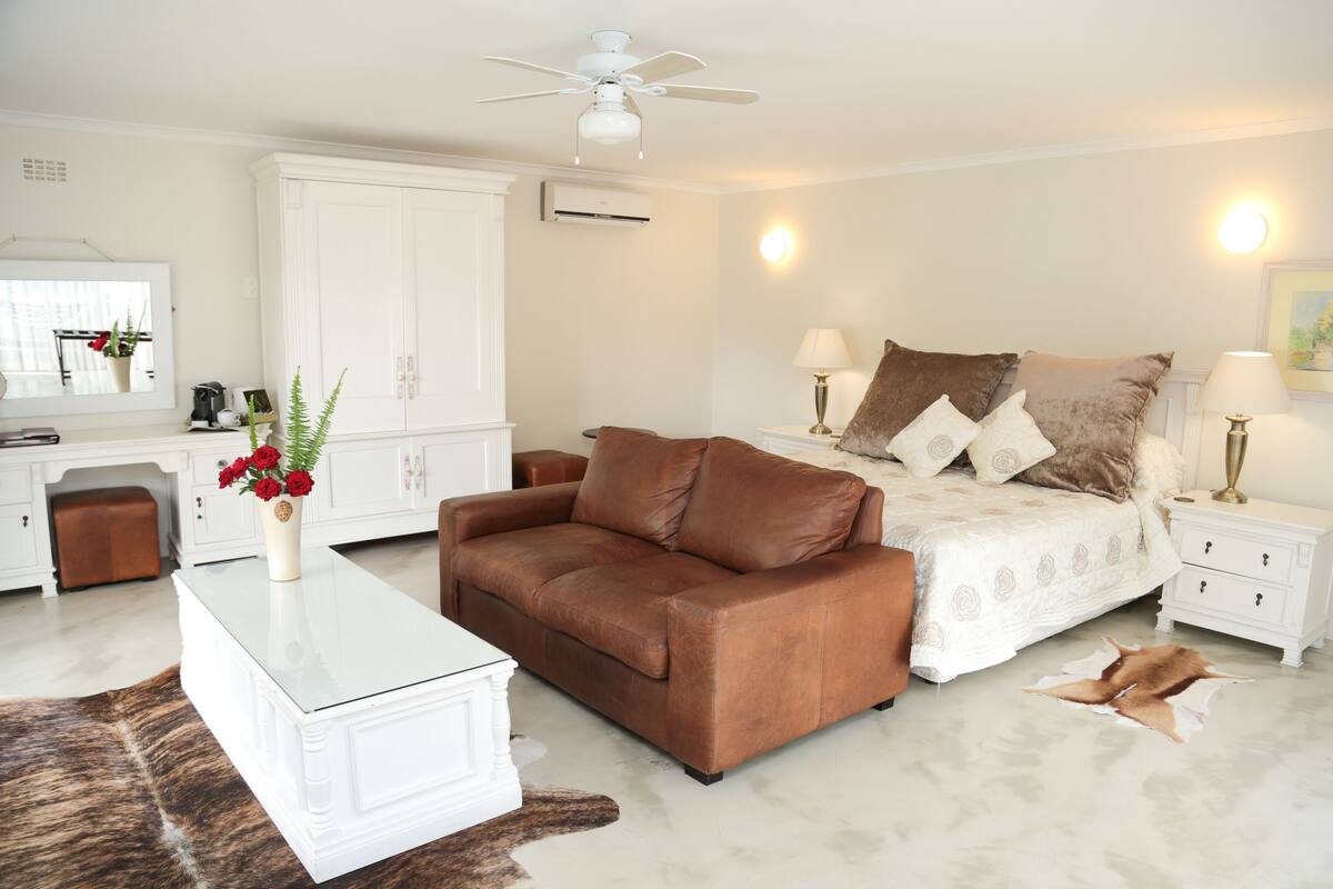 A modern bedroom with a brown leather sofa, a white bed with patterned bedding, and a white nightstand with lamps.