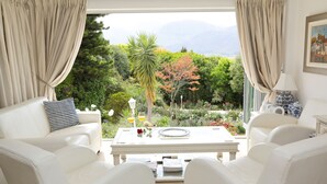 A modern living room with a white sofa set, a dining table with a white tablecloth, and a view of a lush garden and mountains through large windows. - Alba House Bed & Breakfast (Paarl)