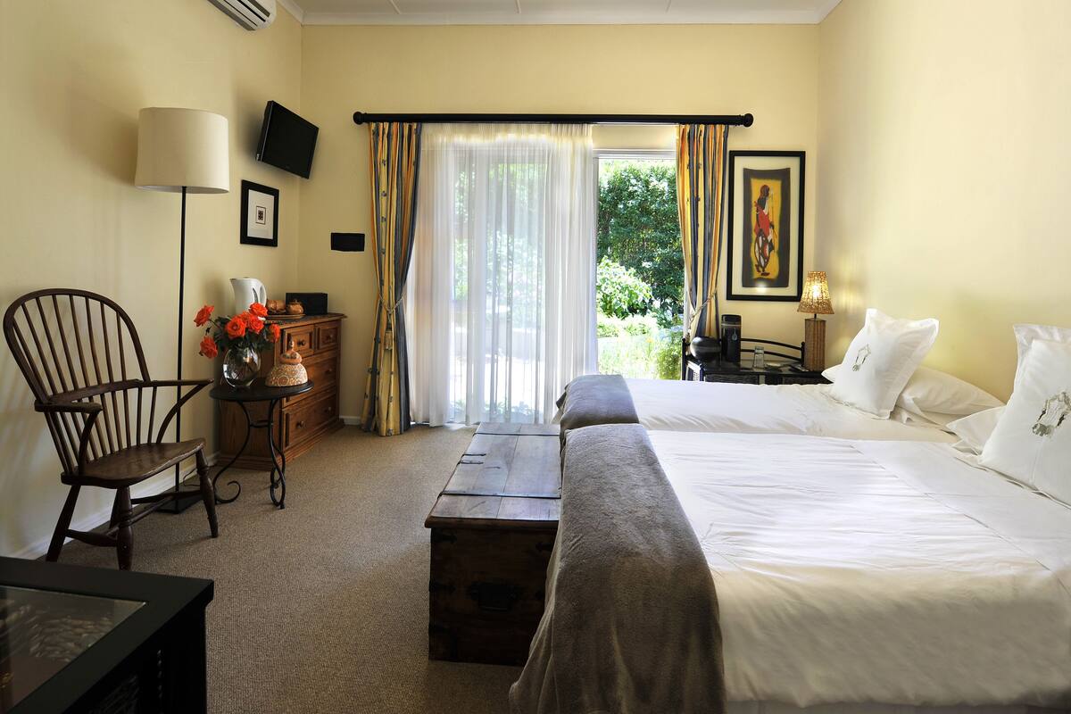 A hotel room with a bed, a chair, a small table with a lamp, a wooden chest, and a view of greenery through the window.
