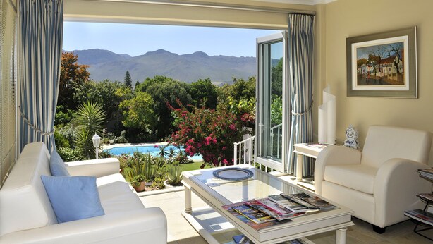 A living room with a sofa, armchairs, a coffee table with magazines, and a view of a pool and mountains. - Alba House Bed & Breakfast (Paarl)