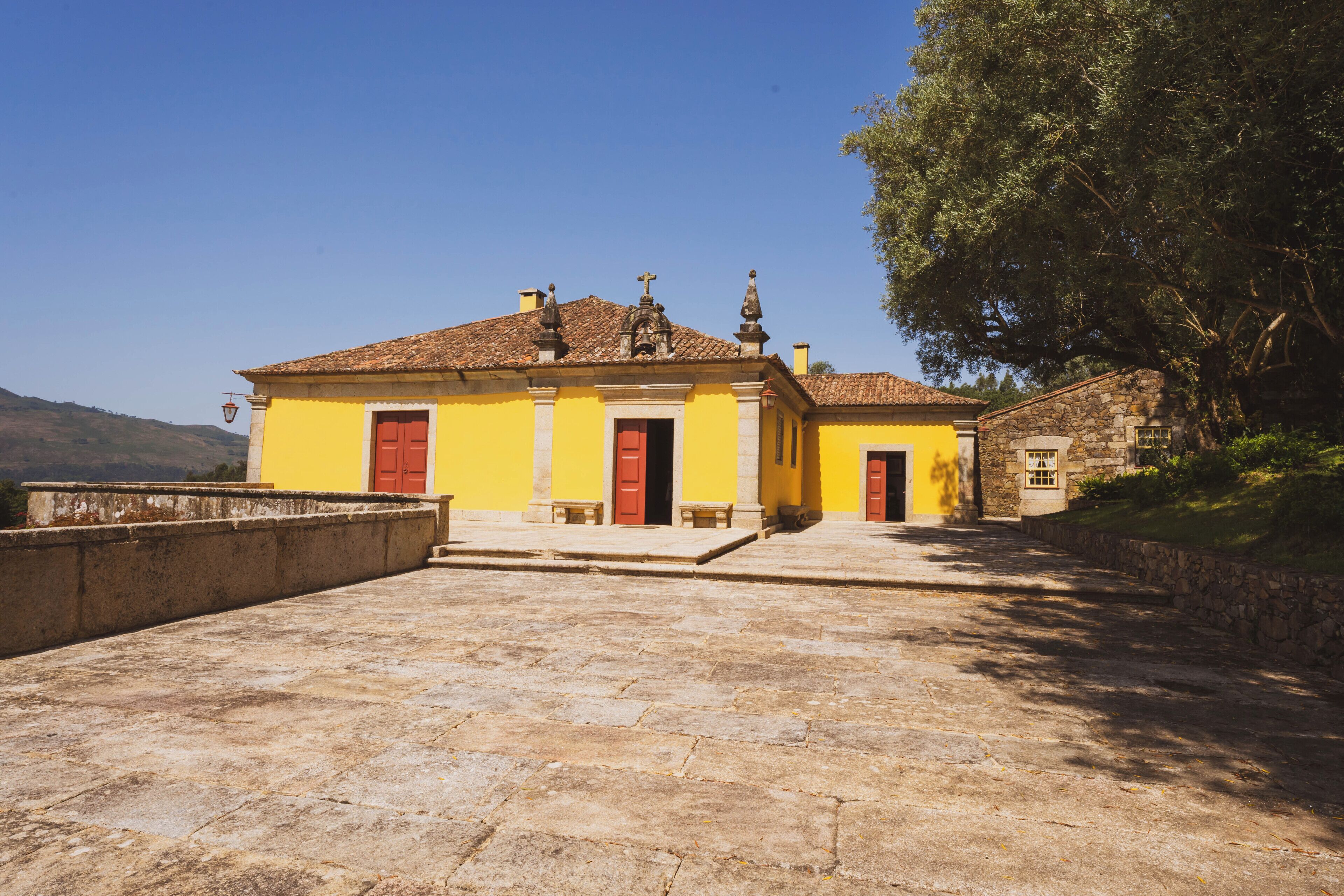 Um edifício amarelo com portas vermelhas, uma parede de pedra e uma grande árvore.