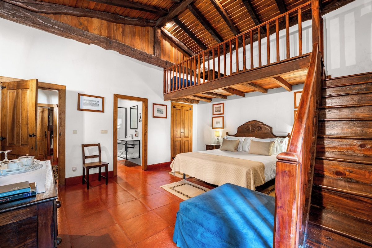 Una habitación con escalera de madera, una cama con cabecera, una silla y una mesa con una bandeja de objetos.