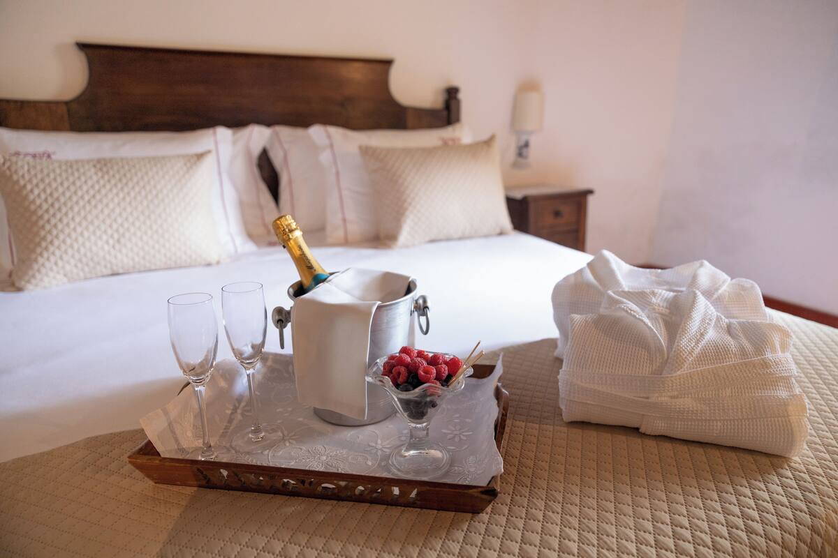 A bed with white linens, a bedside table with a lamp, and a tray with a champagne bottle, glasses, and fruit.