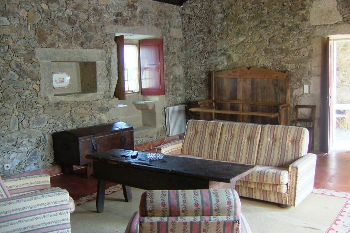 Una habitación con paredes de piedra, un banco de madera, un sillón con cojines y una pequeña mesa.