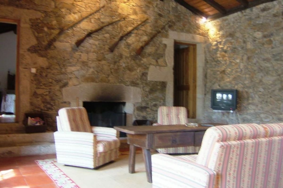 A stone-walled room with a fireplace, striped armchairs, a sofa, and a wooden table.