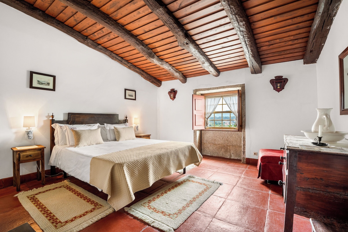 Un dormitorio con techo de madera, una cama con colcha color crema, dos lámparas de noche, una mesita de noche, un banco rojo, un aparador, una ventana con vistas y un cuadro enmarcado en la pared.