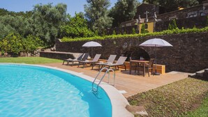 Una piscina con sillones de descanso y sombrillas, rodeada de muros de piedra y follaje.
