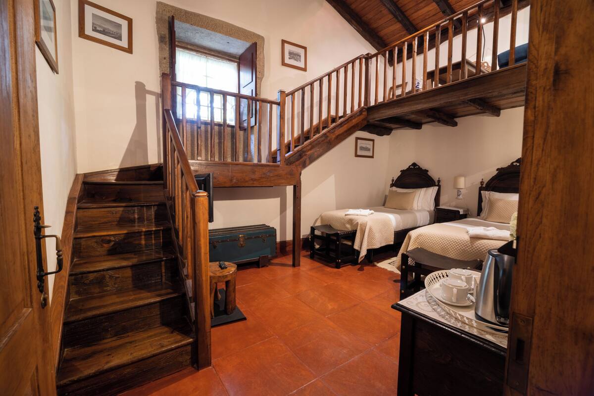 A rustic room with a wooden staircase, a bed with a headboard, a small table with a kettle, and a window with a view.