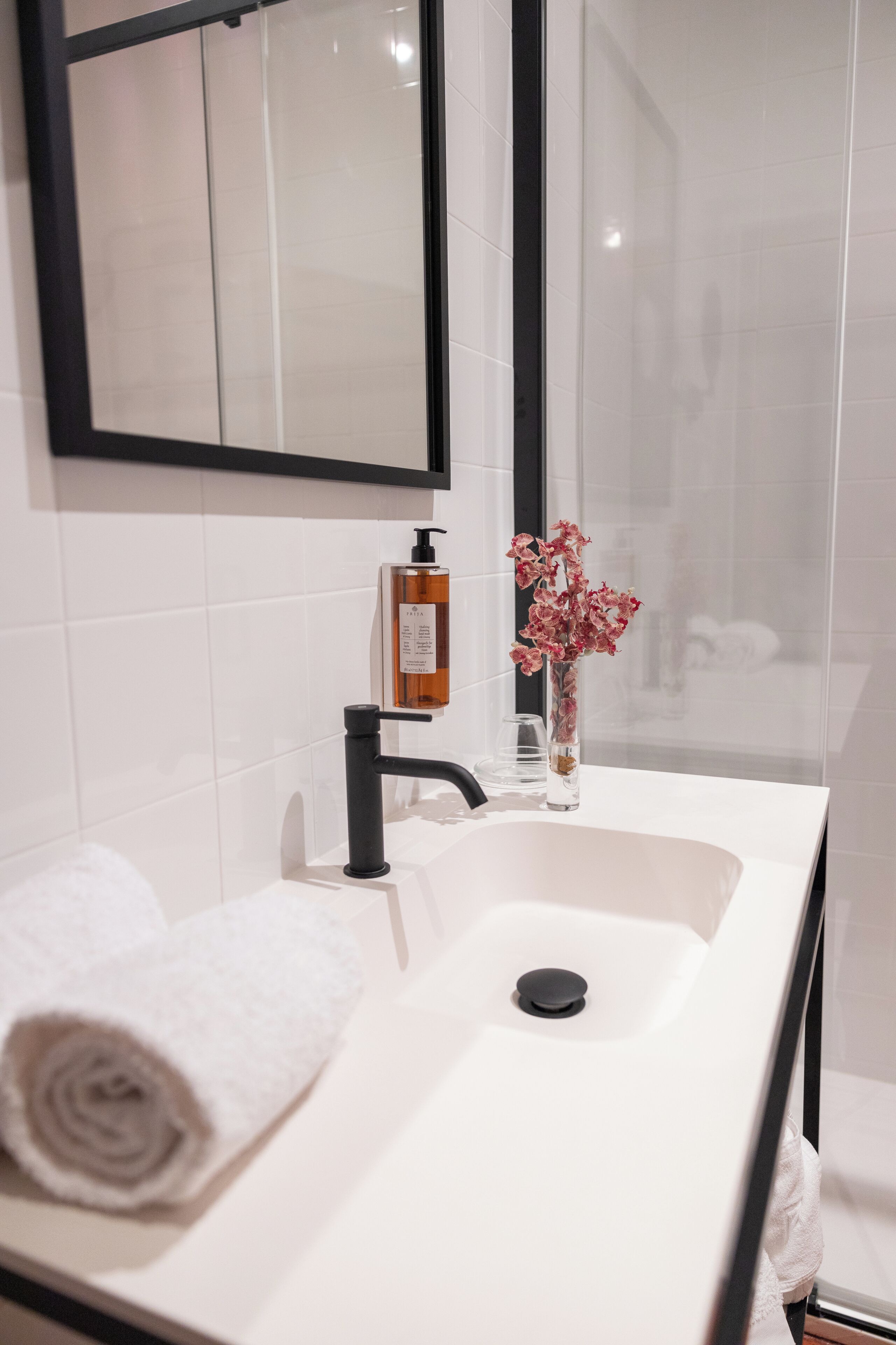 A bathroom with a white sink, black faucet, and a mirror above it. There is a bottle of soap and a vase with flowers on the sink.