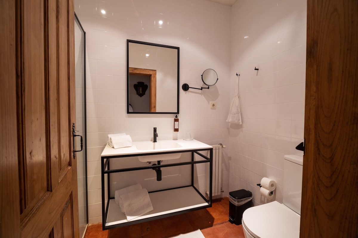 A bathroom with a wooden door, white tiles, a white sink with a black frame, a mirror, a towel, a toilet, and a trash bin.
