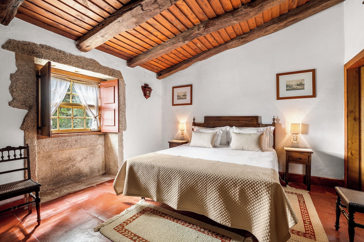 Un dormitorio con techo de madera, una cama con colcha, una mesita de noche, una silla y una ventana con cortinas.