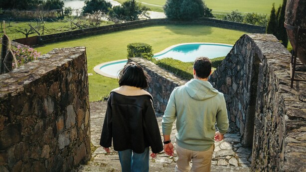 Dos personas caminando sobre escalones de piedra hacia una piscina rodeada de vegetación y colinas.
