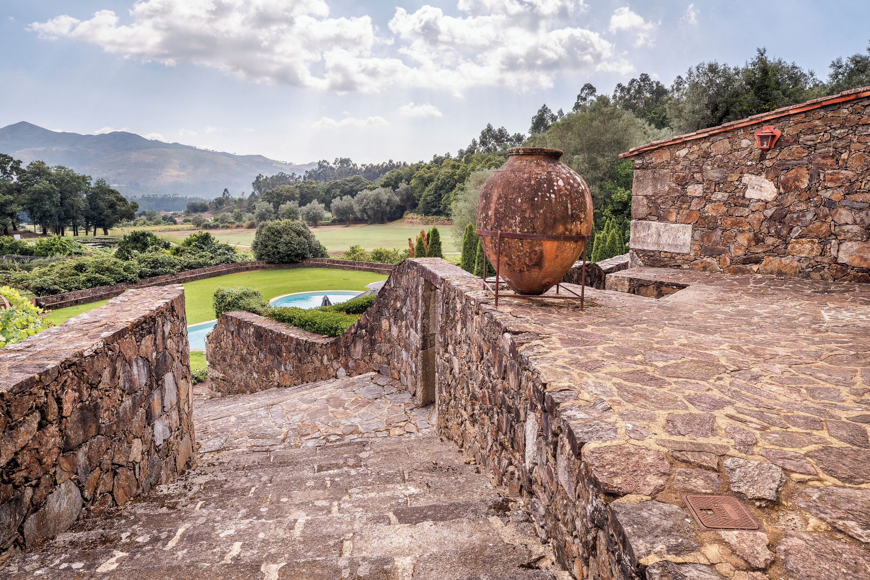 Pátio de pedra com um vaso grande, uma piscina e vista para uma paisagem verde.