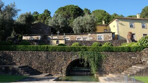 Un puente de piedra con un pequeño arco que da a una zona de piscina, rodeada de abundante vegetación y edificios históricos.