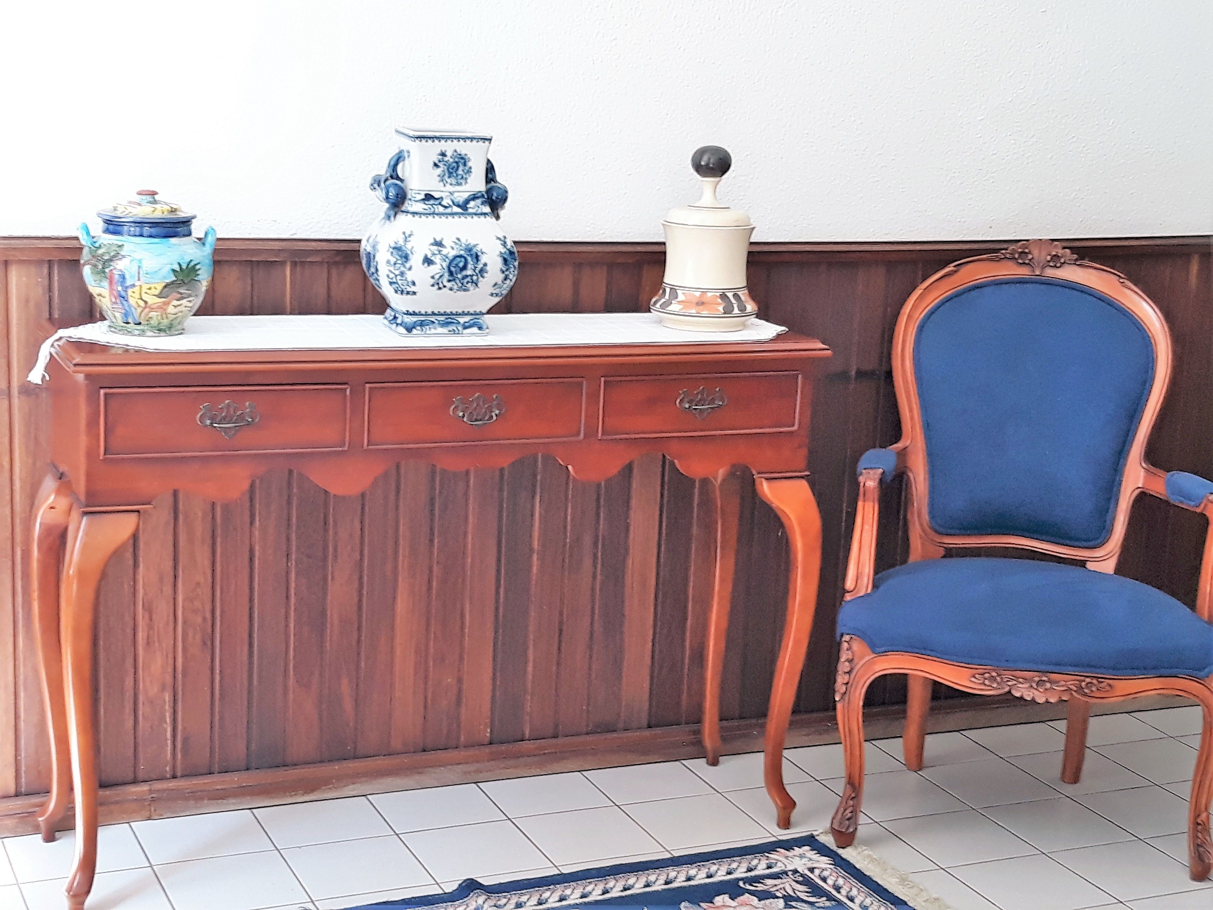Una scrivania in legno con tre vasi decorativi e un saliera, una poltrona rivestita in blu e un tappeto a motivi su un pavimento piastrellato.