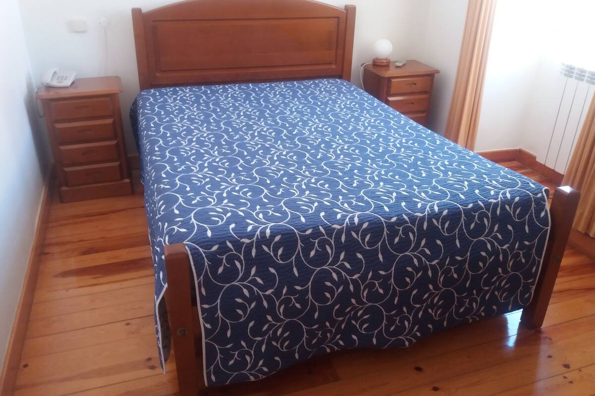 A bedroom with a wooden bed, a nightstand, a bedside table, and a wall socket.