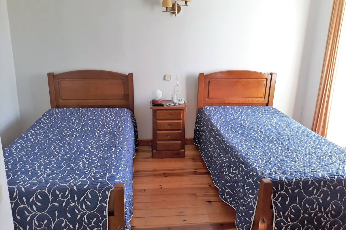 A room with two wooden beds, each with a blue patterned bedspread, a wooden nightstand with a lamp, and a wall-mounted light fixture.