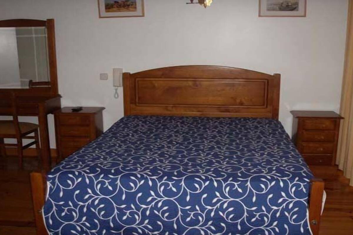 A bedroom with a wooden bed, a nightstand, a dresser, a mirror, and a wall-mounted light fixture.