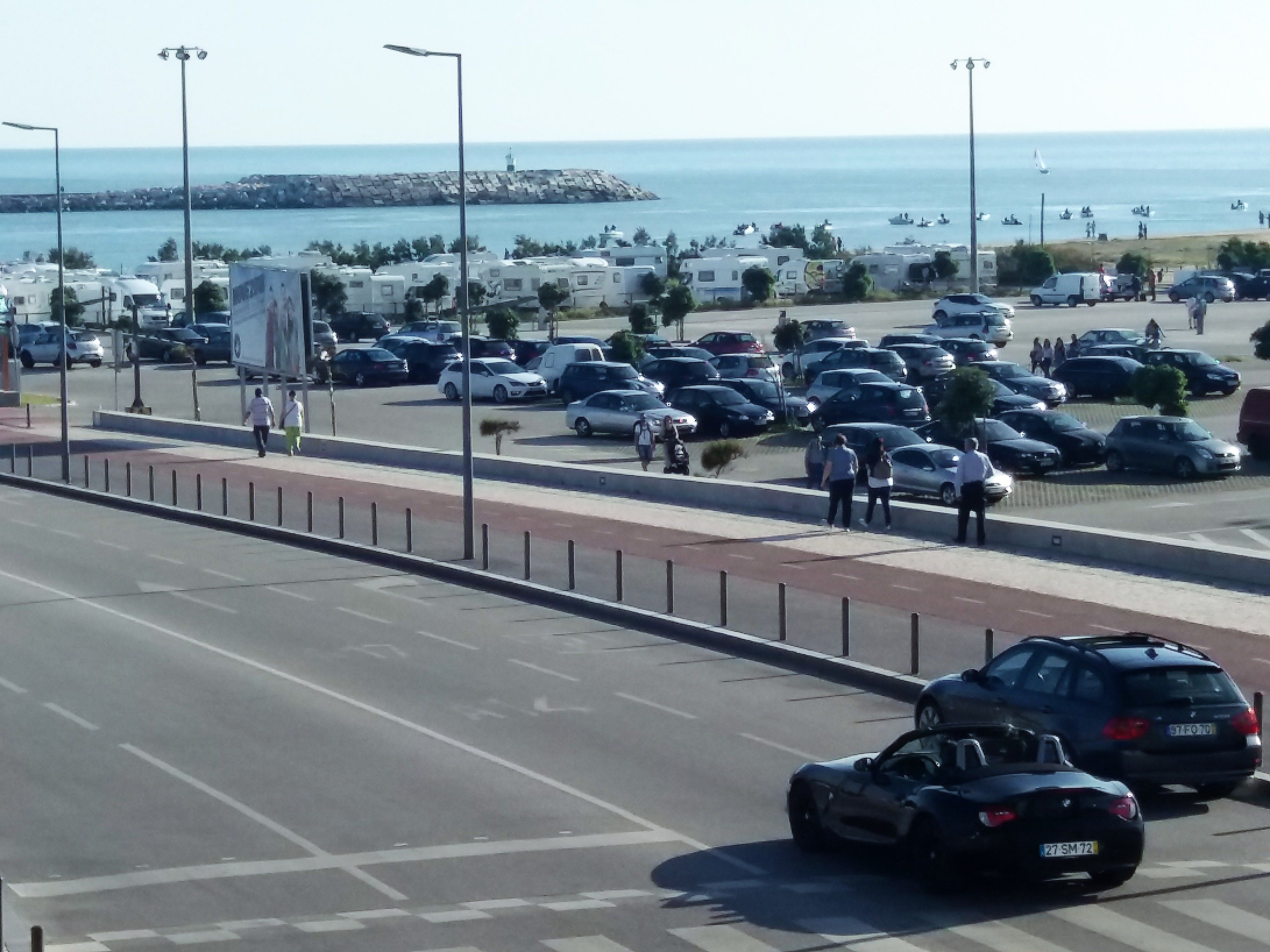 A parking lot with RVs and cars, a road with a pedestrian crossing, and a view of the sea and buildings in the background.