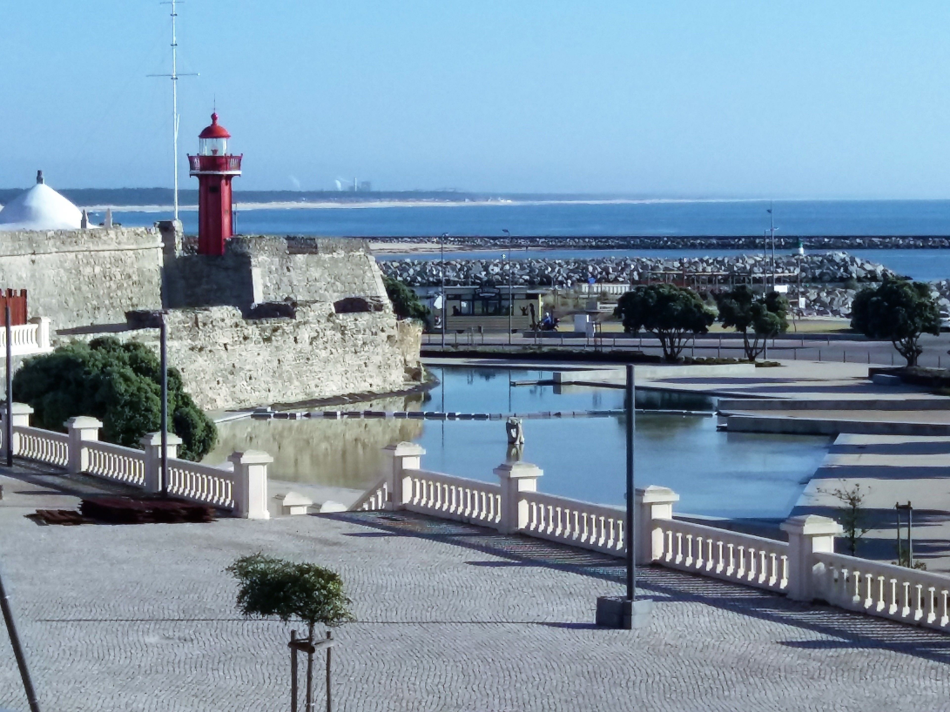 A coastal promenade with a red lighthouse, stone walls, and a calm sea.