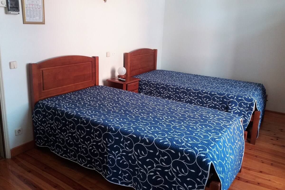 A room with two wooden beds, a small bedside table with a lamp, and a framed notice on the wall.