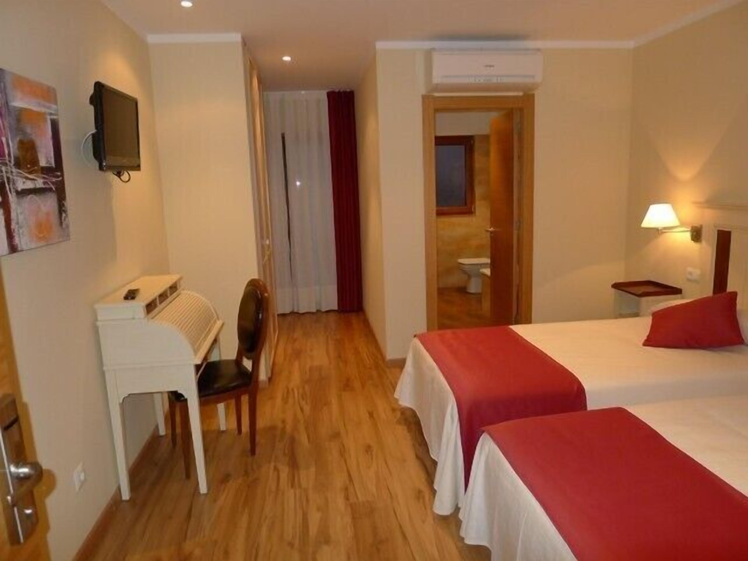 Habitación de hotel con dos camas, un escritorio, una silla, televisión y baño visible a través de una puerta abierta.