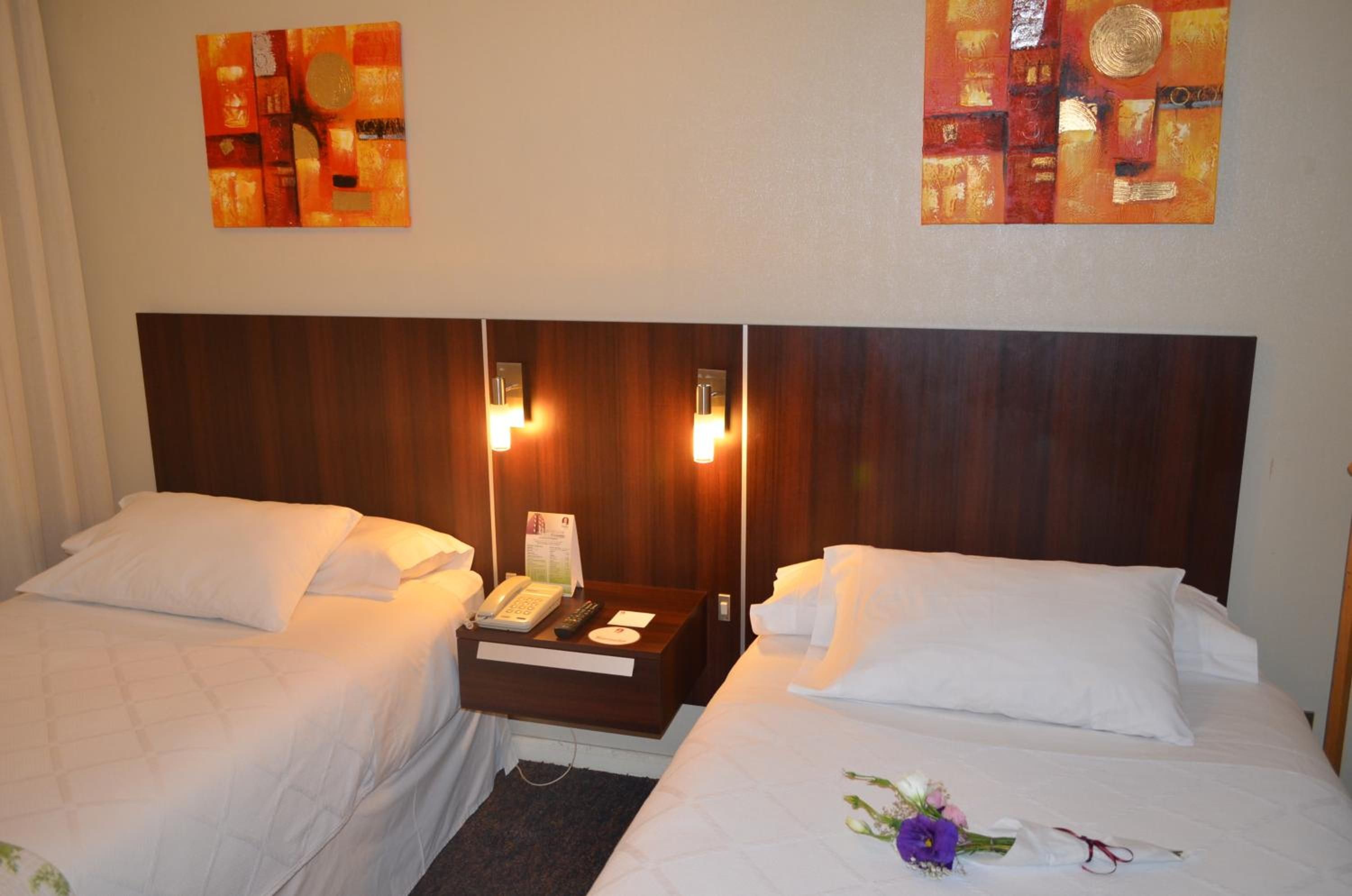 A hotel room with two beds, a nightstand with a phone and remote, and artwork on the walls.
