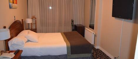 Una habitación de hotel con una cama, un escritorio con teléfono, una lámpara, un televisor y una ventana con cortinas.