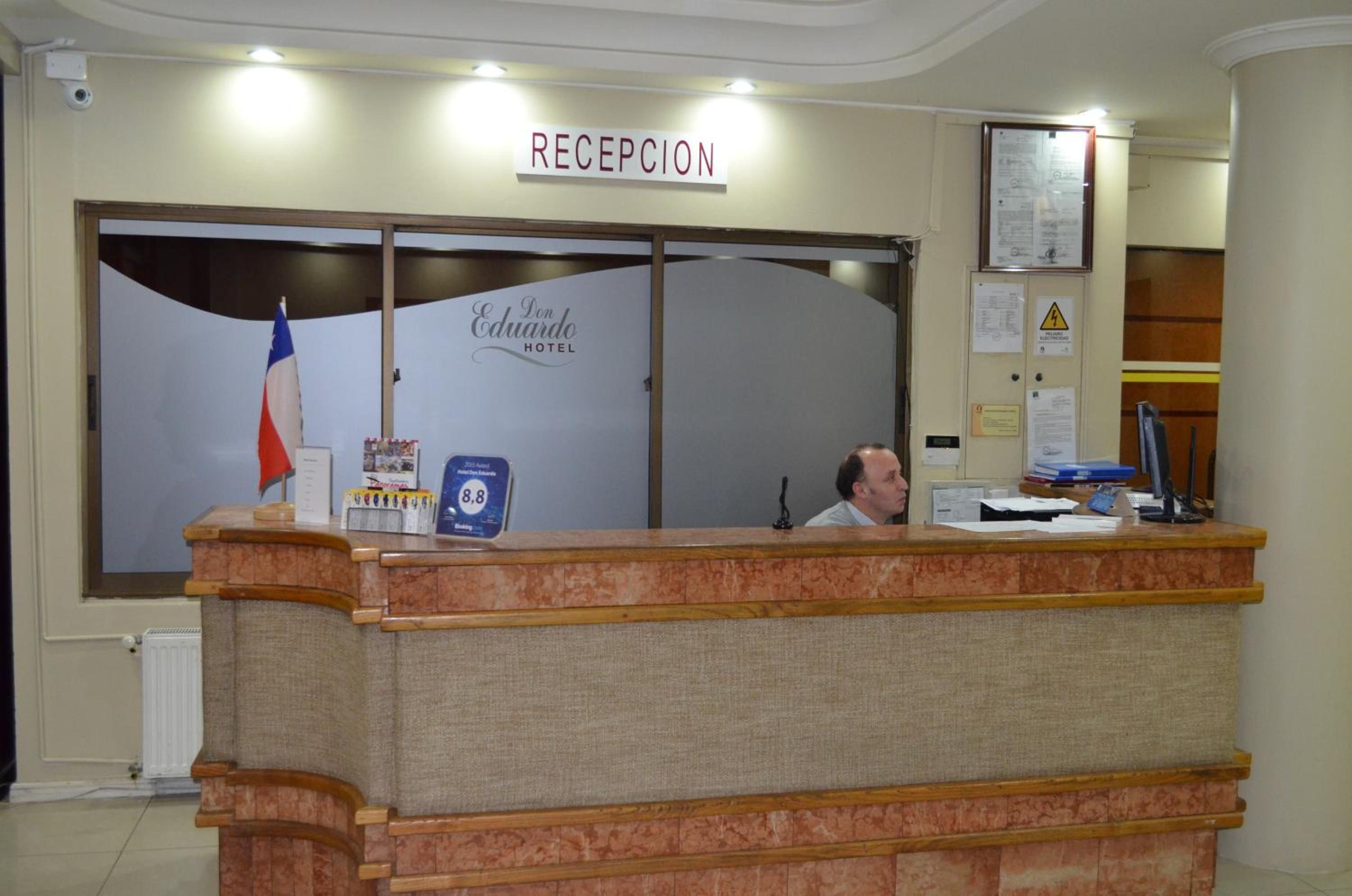 Un comptoir d’accueil surmonté d’une enseigne indiquant « RÉCEPTION ». Derrière le comptoir, on aperçoit une fenêtre avec un drapeau et une enseigne indiquant « HÔTEL Don Eduardo ».