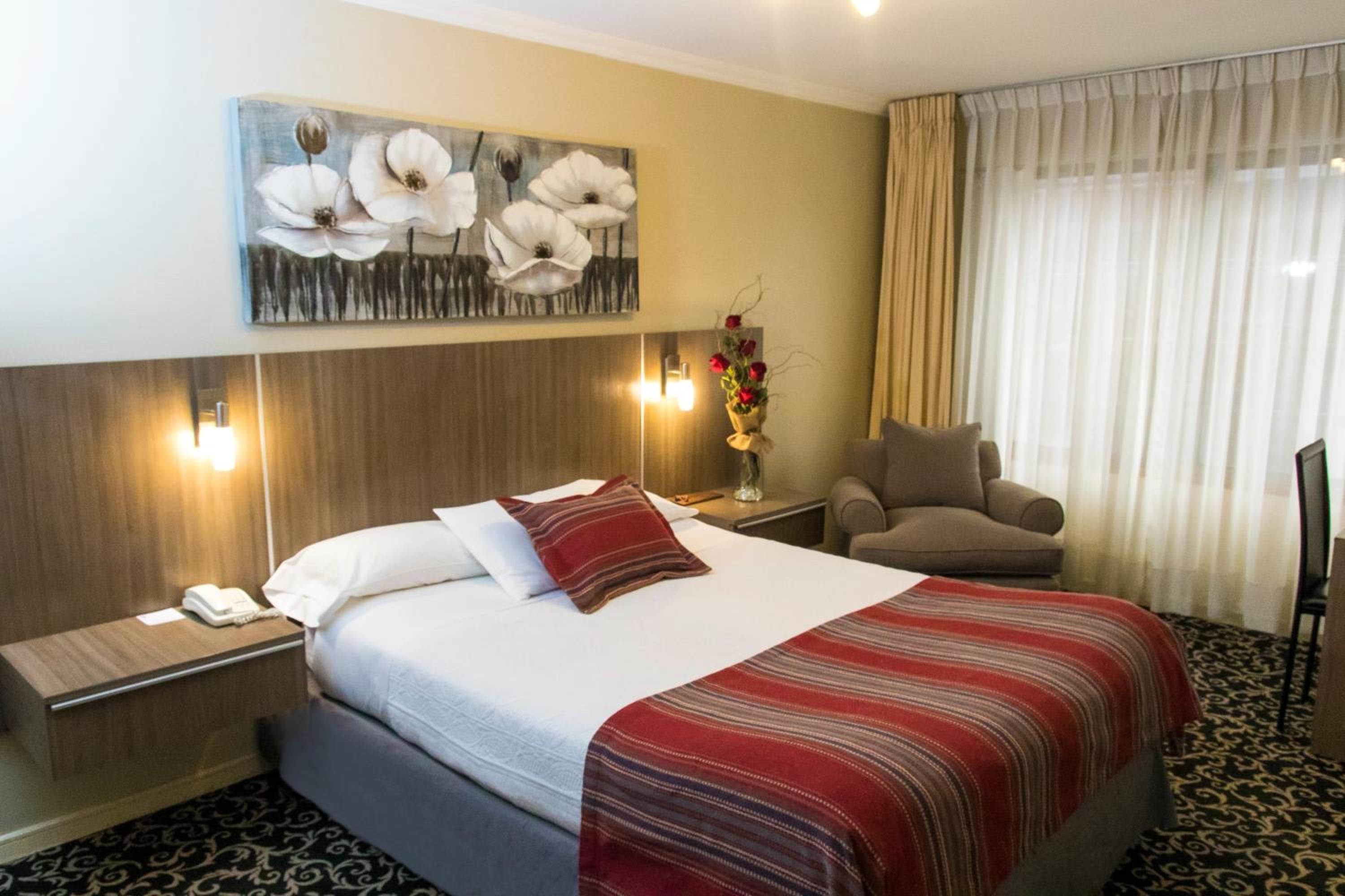Una habitación de hotel con cama, una silla, una mesita de noche, un jarrón con flores y un cuadro en la pared.
