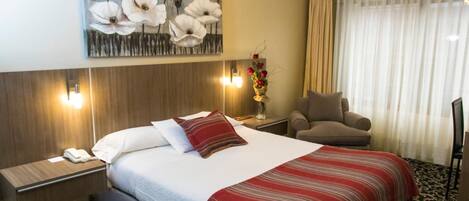 Una habitación de hotel con cama, una silla, una mesita de noche, un jarrón con flores y un cuadro en la pared.