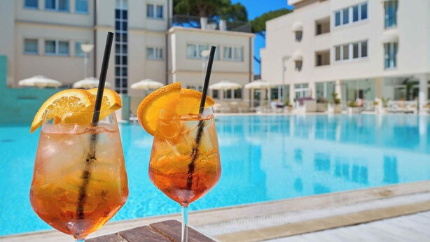 Deux boissons, décorées de tranches d’orange et servies avec des pailles, sont posées au bord d’une piscine, avec un bâtiment visible en arrière-plan.
