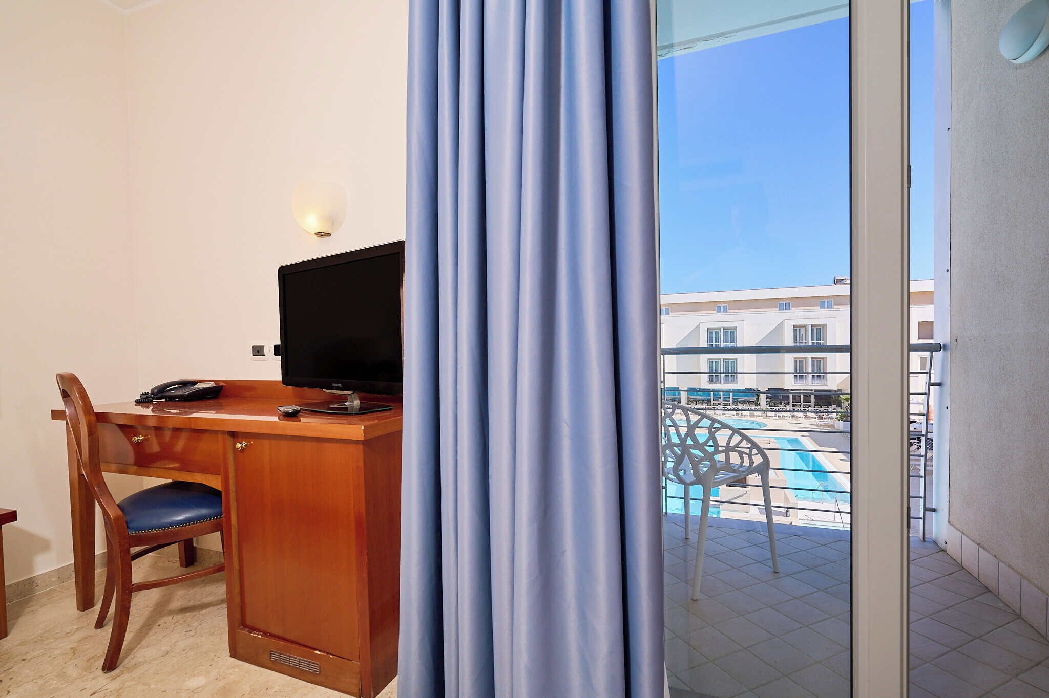 Een hotelkamer met een bureau, stoel, televisie en uitzicht op een zwembad en gebouwen.
