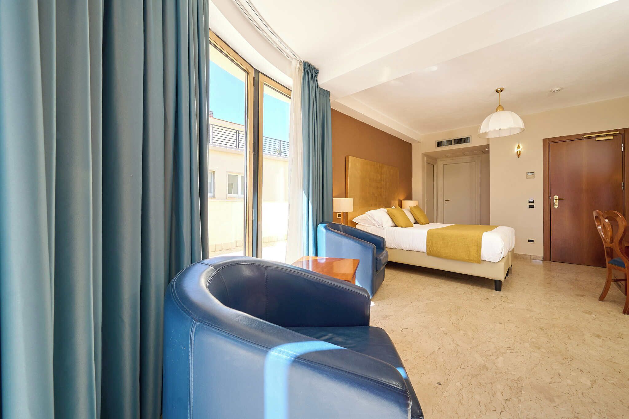 Een hotelkamer met een bed, twee blauwe stoelen, een tafel, een lamp en uitzicht op buiten.