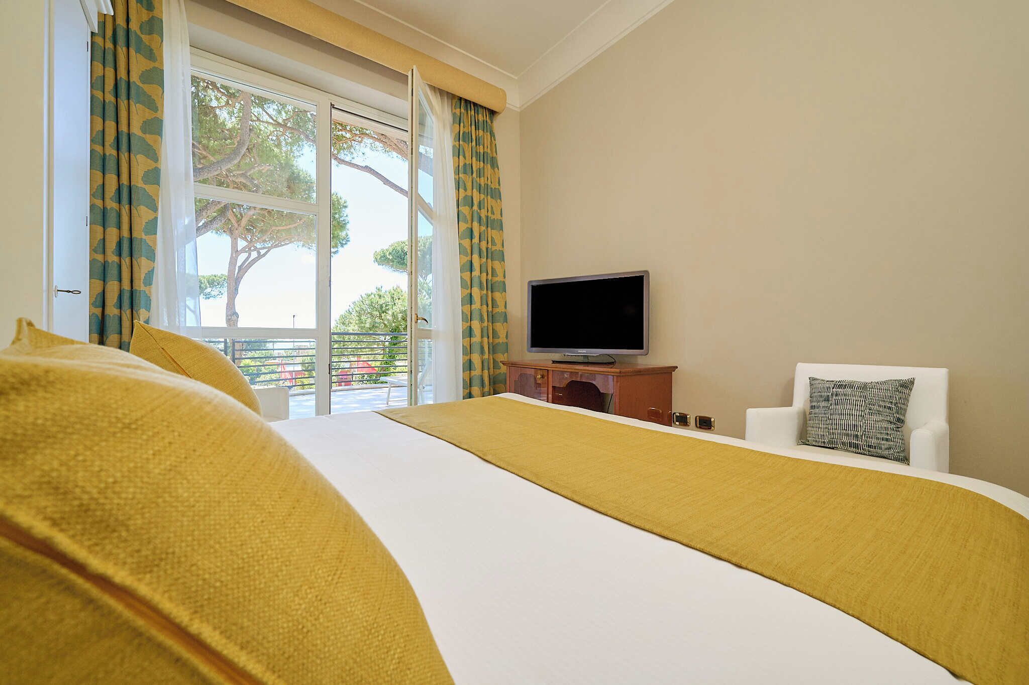 A hotel room with a bed, a television on a wooden stand, a white armchair, and a view of the outdoors through a window with patterned curtains.
