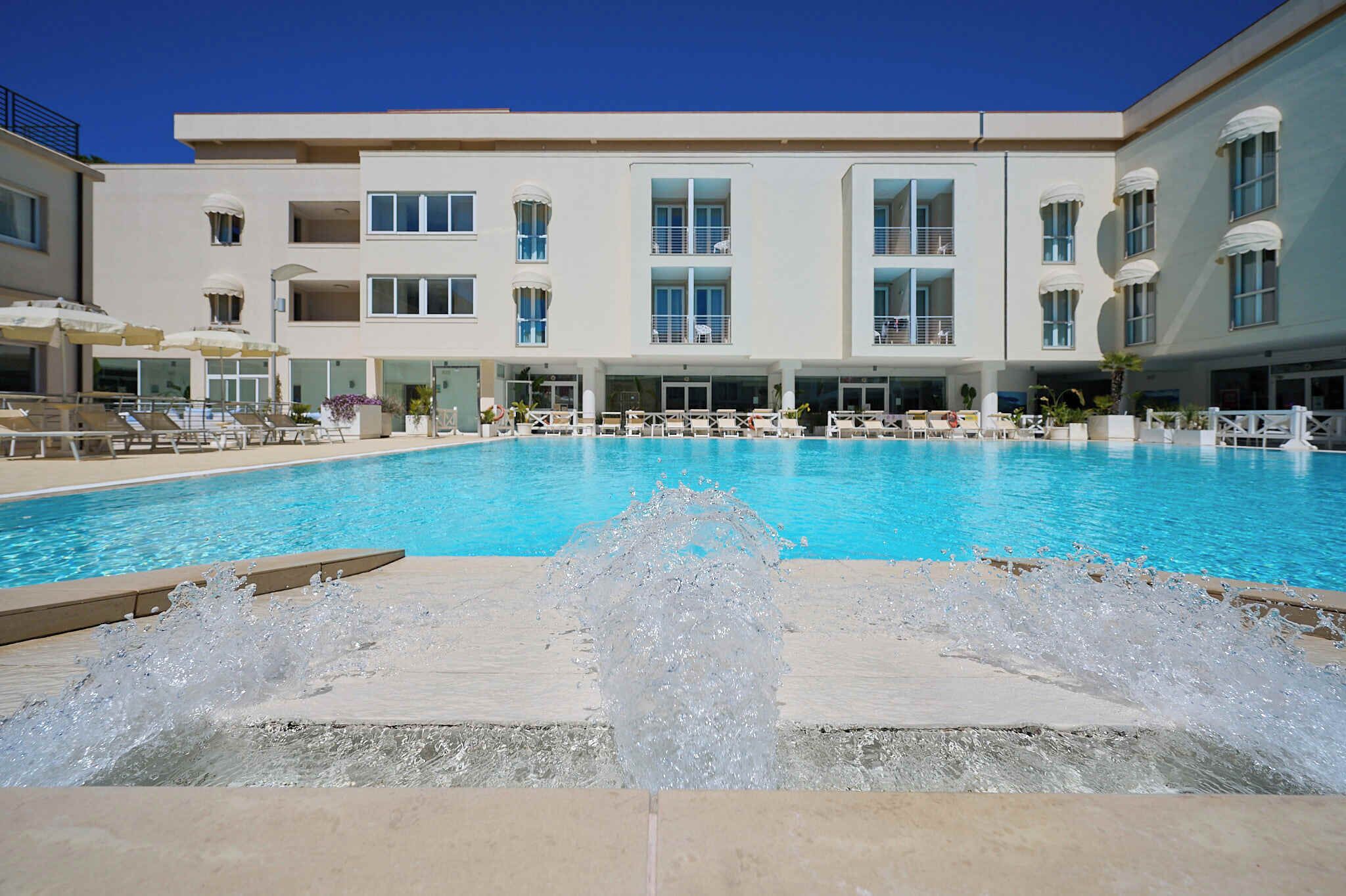 A swimming pool with a water fountain feature, surrounded by lounge chairs and umbrellas, with a multi-story building in the background.