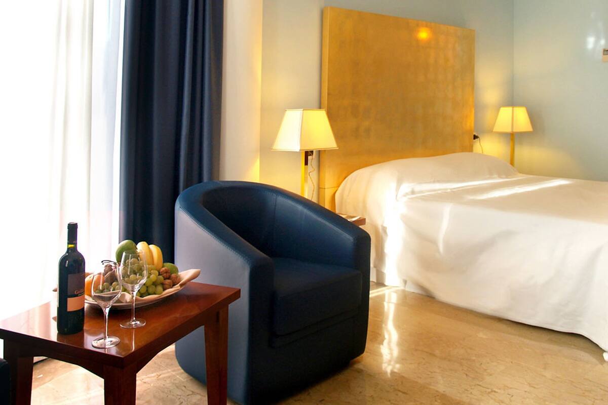 Ein Hotelzimmer mit einem Bett, einem blauen Sessel, einem Holztisch mit Flasche und Gläsern sowie einer Obstschale.
