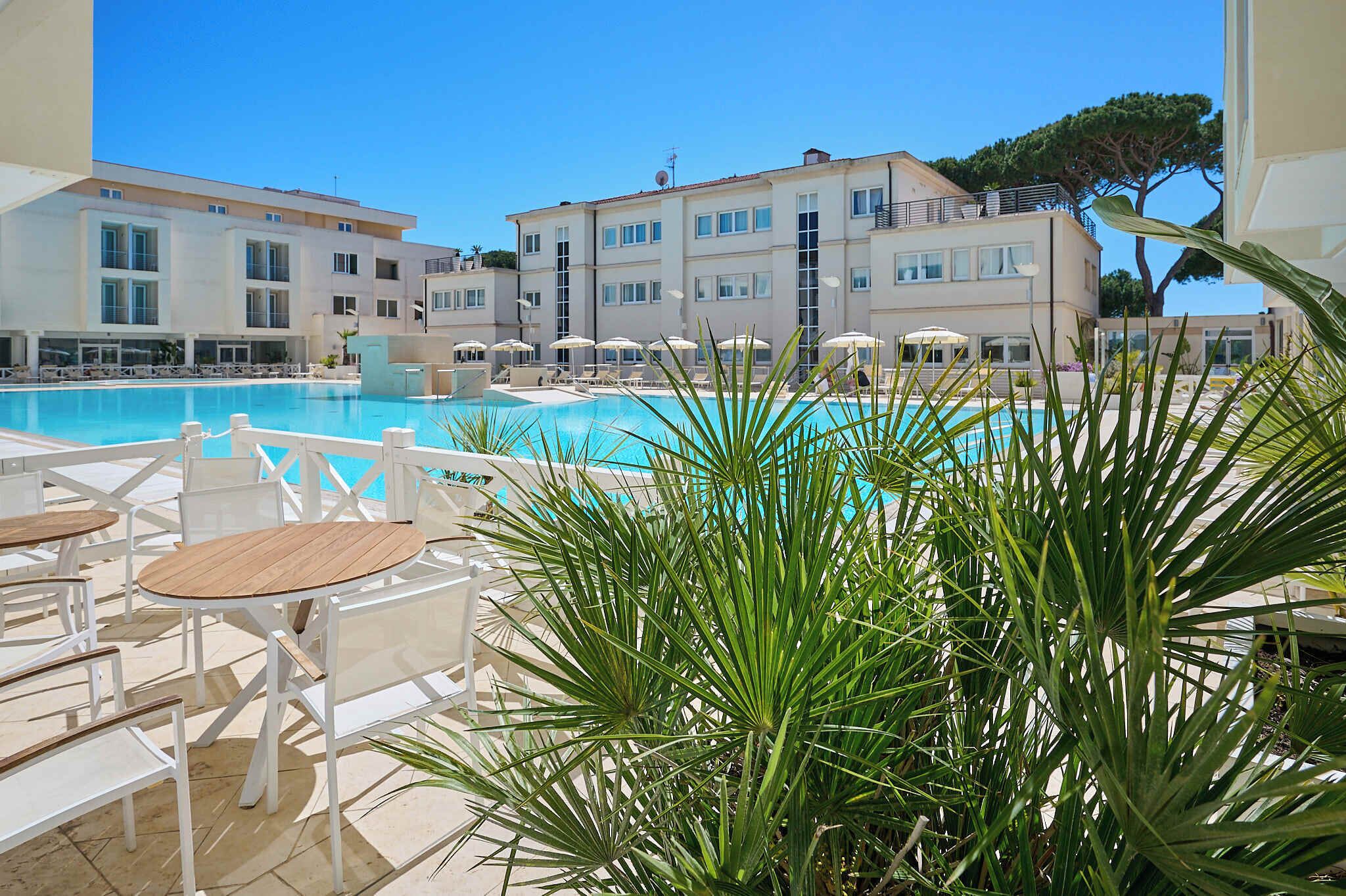 A swimming pool with sun loungers and umbrellas, surrounded by buildings with balconies and a tree.