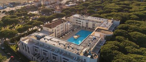 Un edificio grande con piscina en la azotea, rodeado de abundante vegetación y vistas al mar.