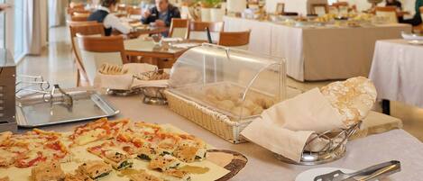 A buffet with various food items, including pizza and pastries, in a dining area with tables and chairs.