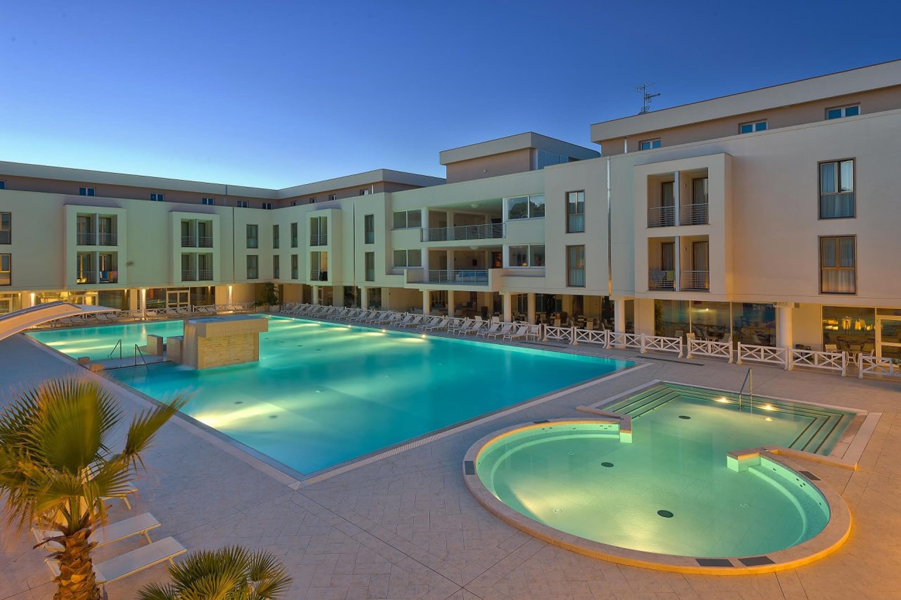 A modern hotel with a large outdoor swimming pool, multiple balconies, and a well-lit terrace area.