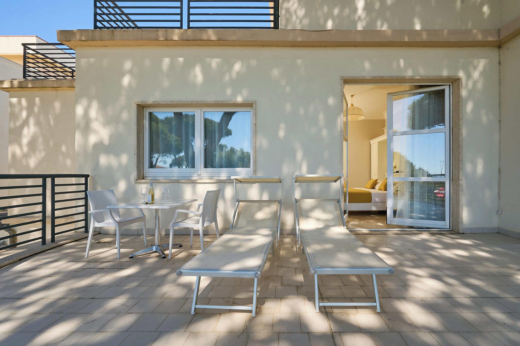Eine Terrasse mit Tisch und Stühlen, eine Bank sowie eine Glastür, die in einen Raum mit einem Bett und einer Lampe führt.