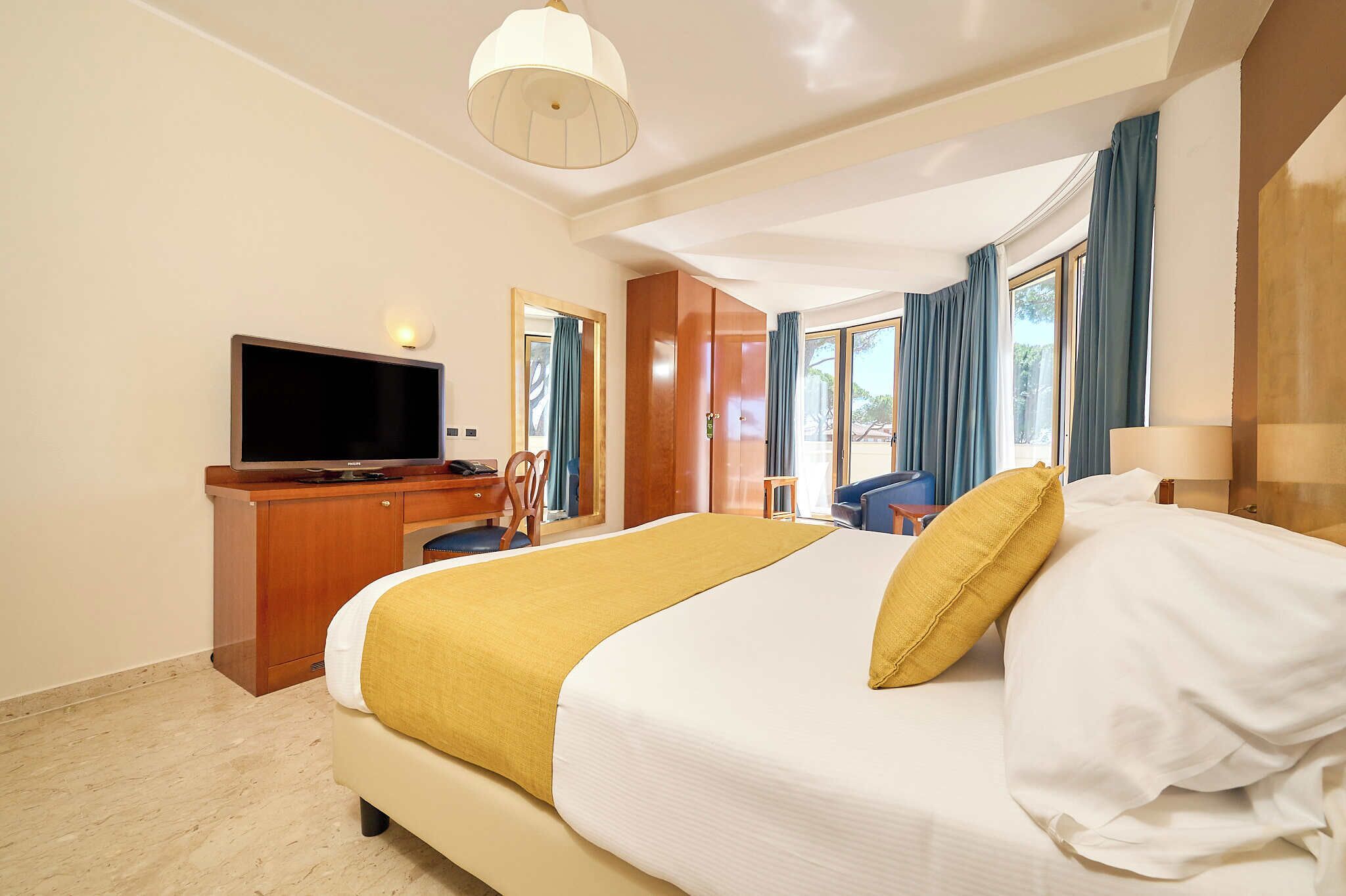A hotel room with a bed, a desk with a television, a chair, a lamp, and large windows with curtains.