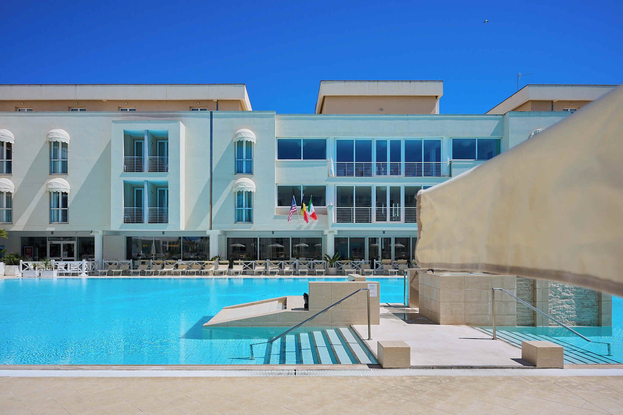 A swimming pool with lounge chairs, a flag, and a building with large windows and balconies.