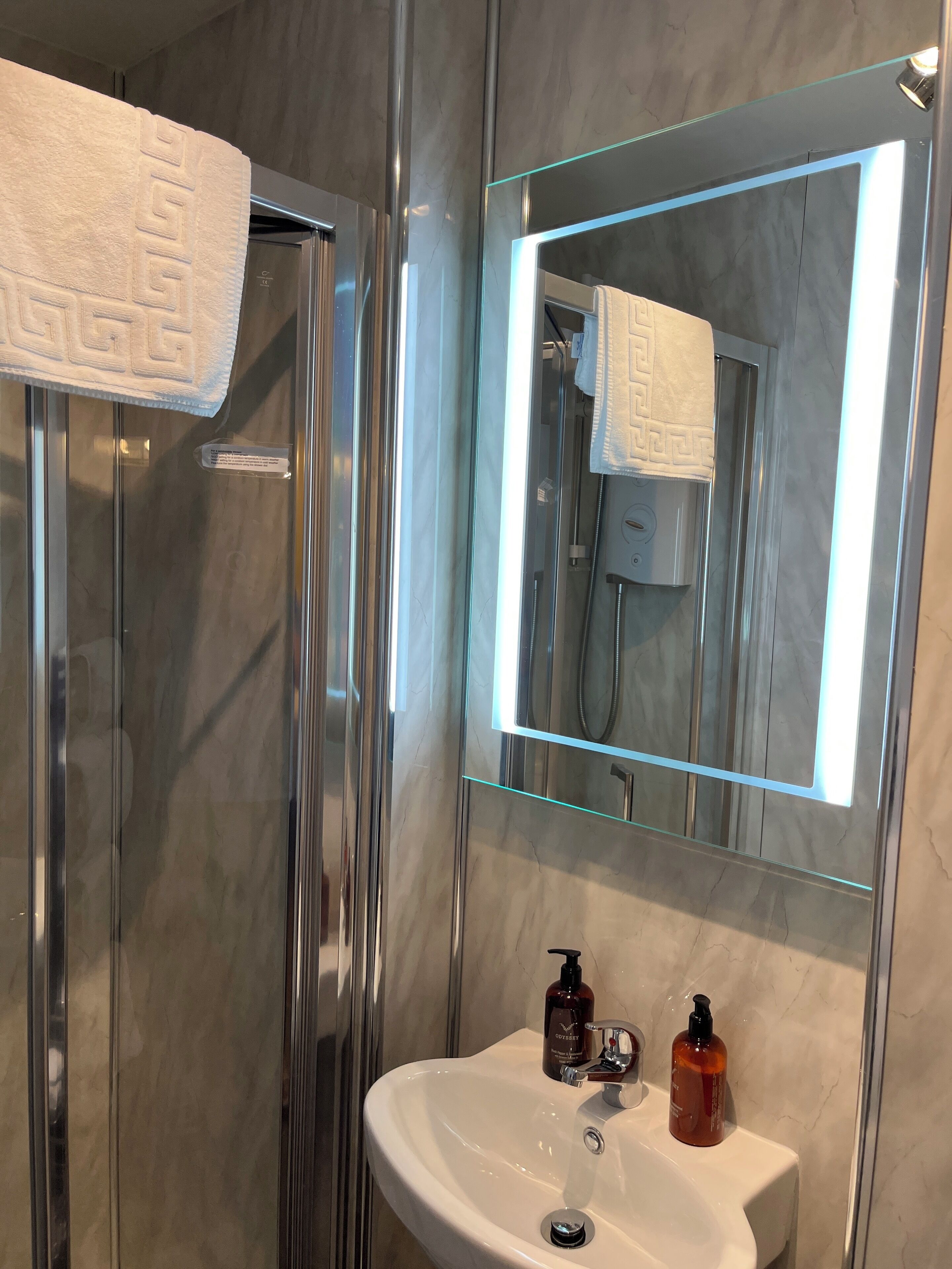 Et badeværelse med brusekabine i glas, en vask med et spejl over og toiletartikler på vasken.